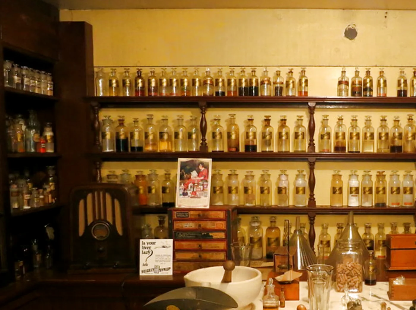 Regalwand einer alten Apotheke: Reihen beschrifteter Glasflaschen über einem Marmortisch mit Mörser und Waage
